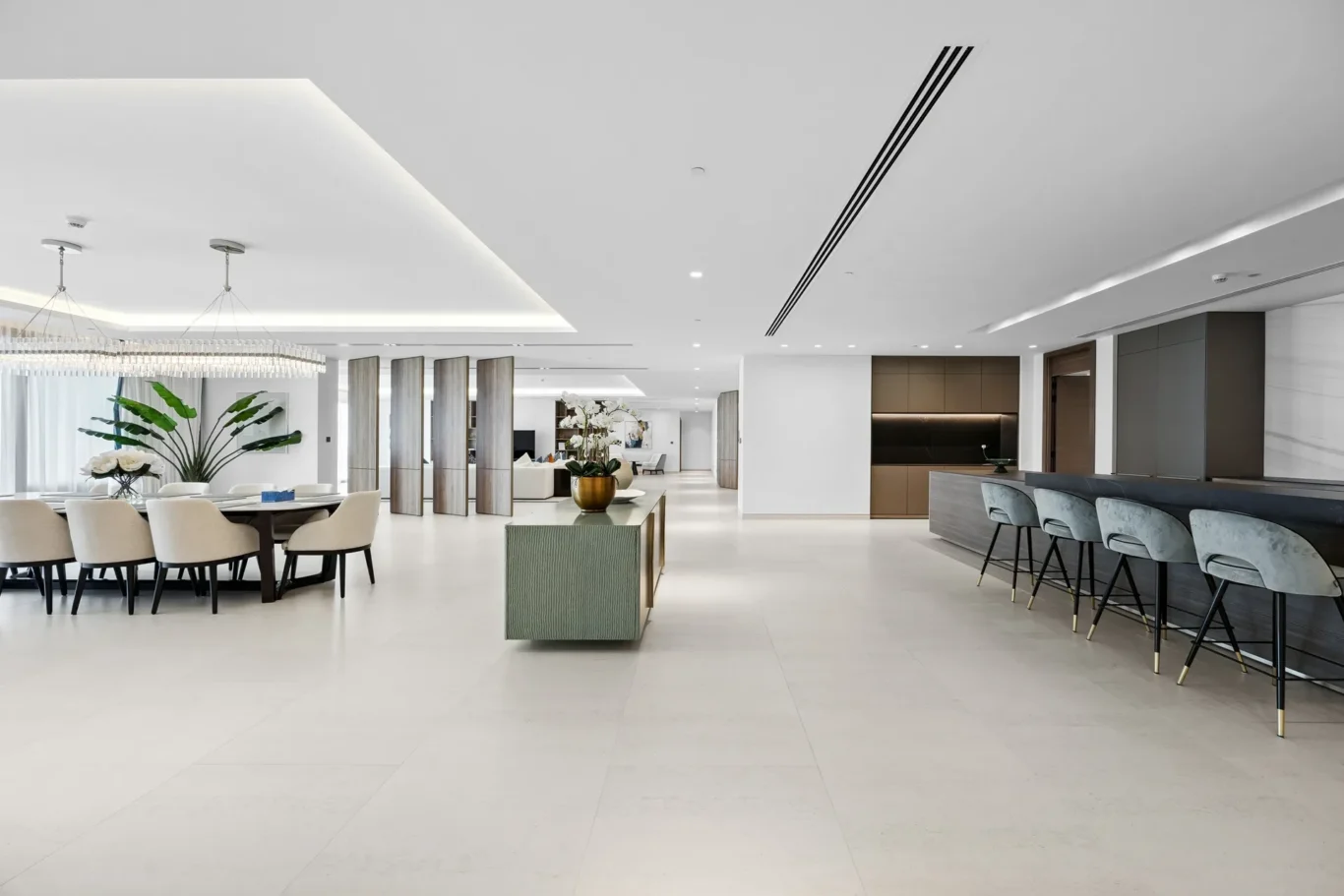 Spacious modern lobby with a dining area on the left, plants, and a sleek bar along the right wall; neutral tones throughout.