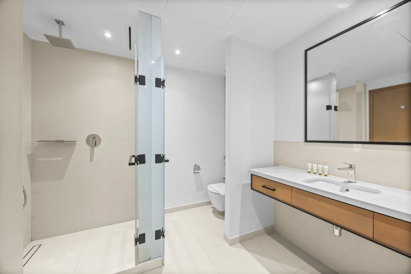 Modern bathroom with glass shower enclosure, white tiles, double-sink vanity with wooden drawers, and a large framed mirror.
