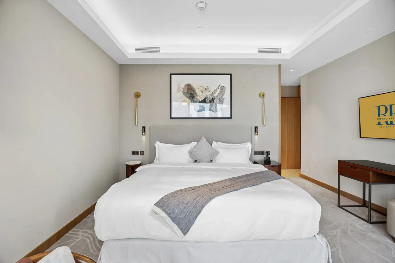 Hotel room with a large white bed, gray headboard, and matching bedding in neutral tones; framed art above the bed and gold wall lamps on each side.