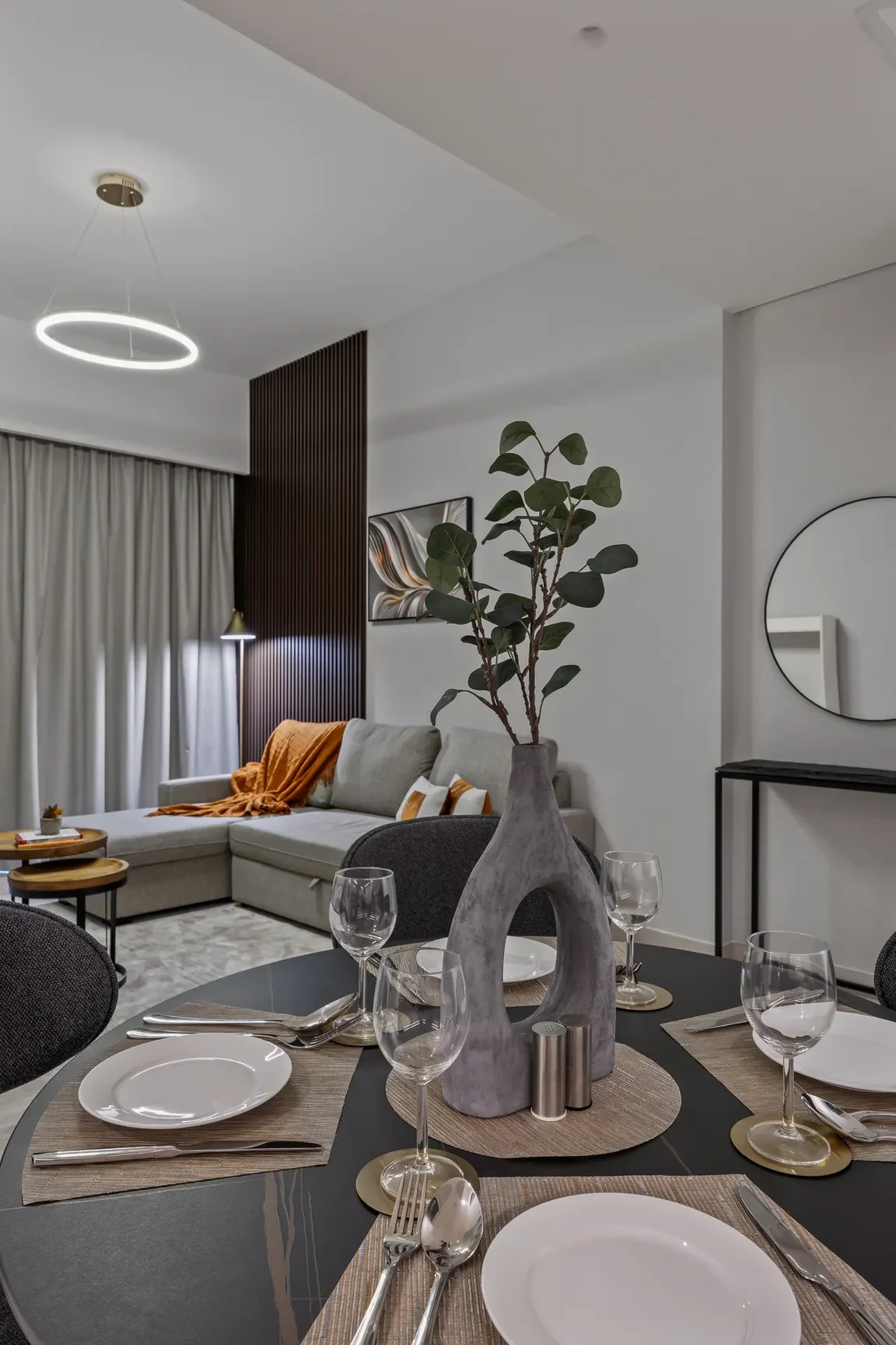 Round dining table set for four with white plates, silverware, and a gray centerpiece vase in a modern living-dining space.