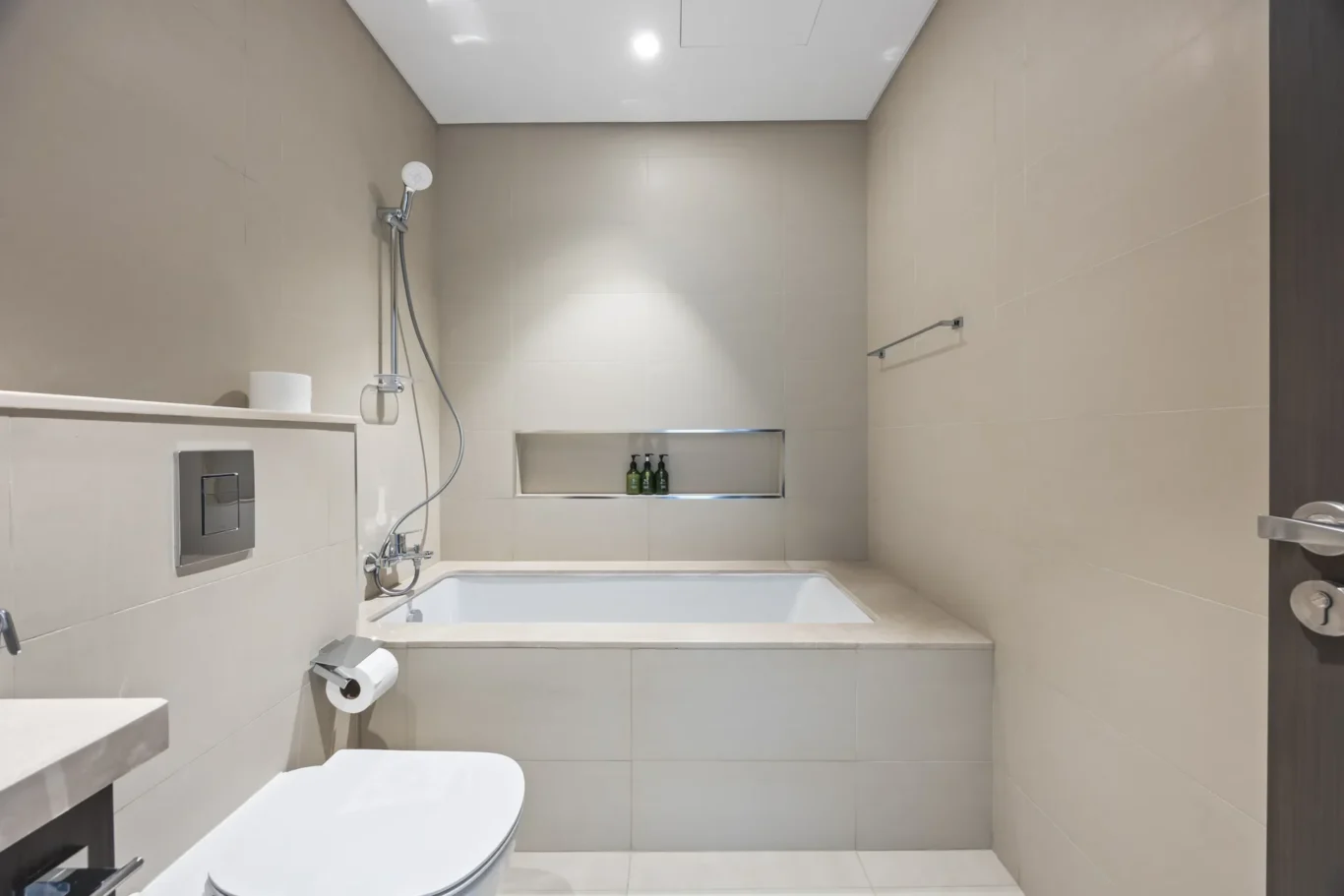 Modern beige-tiled bathroom with a built-in shower niche containing three bottles above a white bathtub.