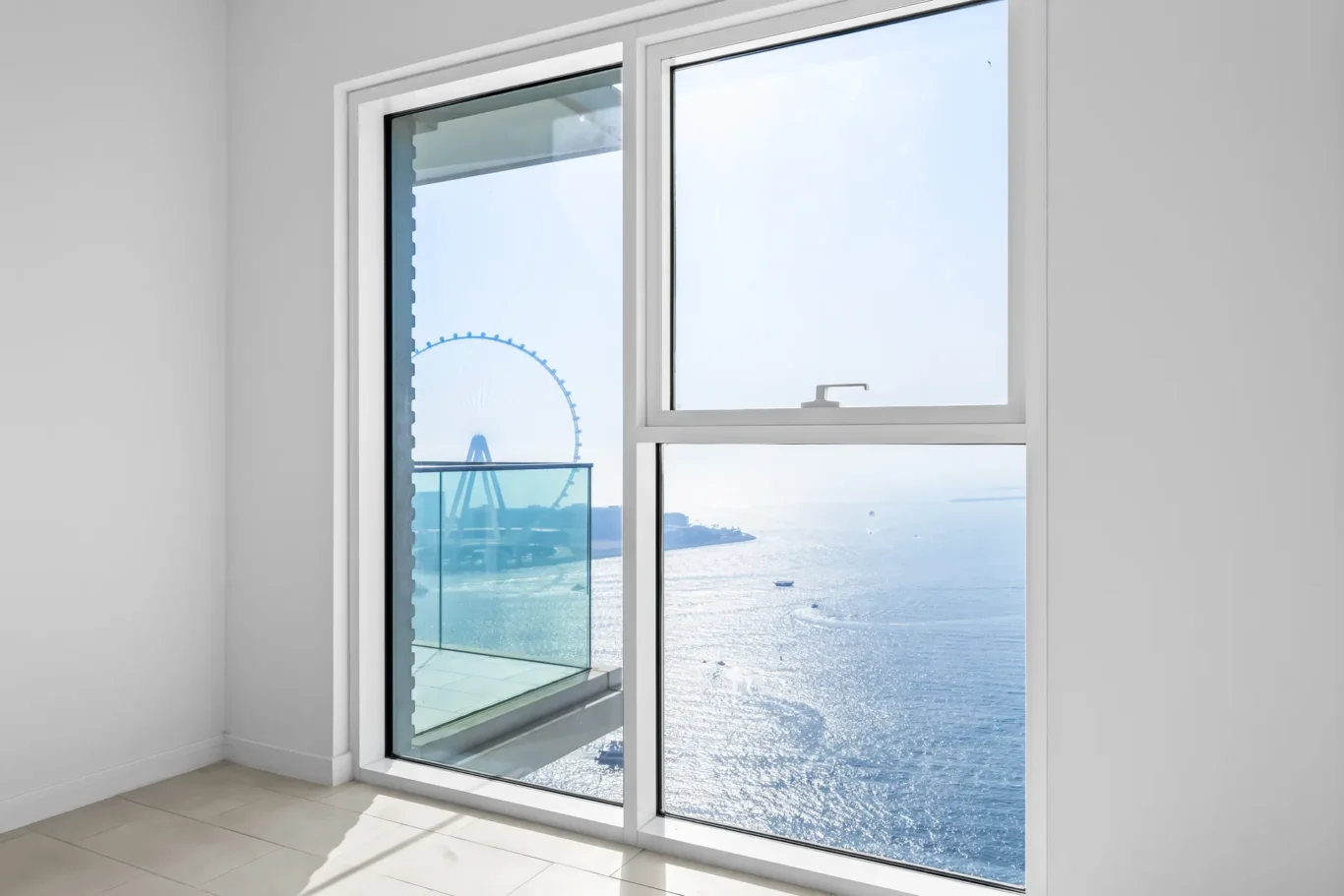 Bright room with a large white-framed window; outside is a seaside view with a ferris wheel and glass balcony railing.