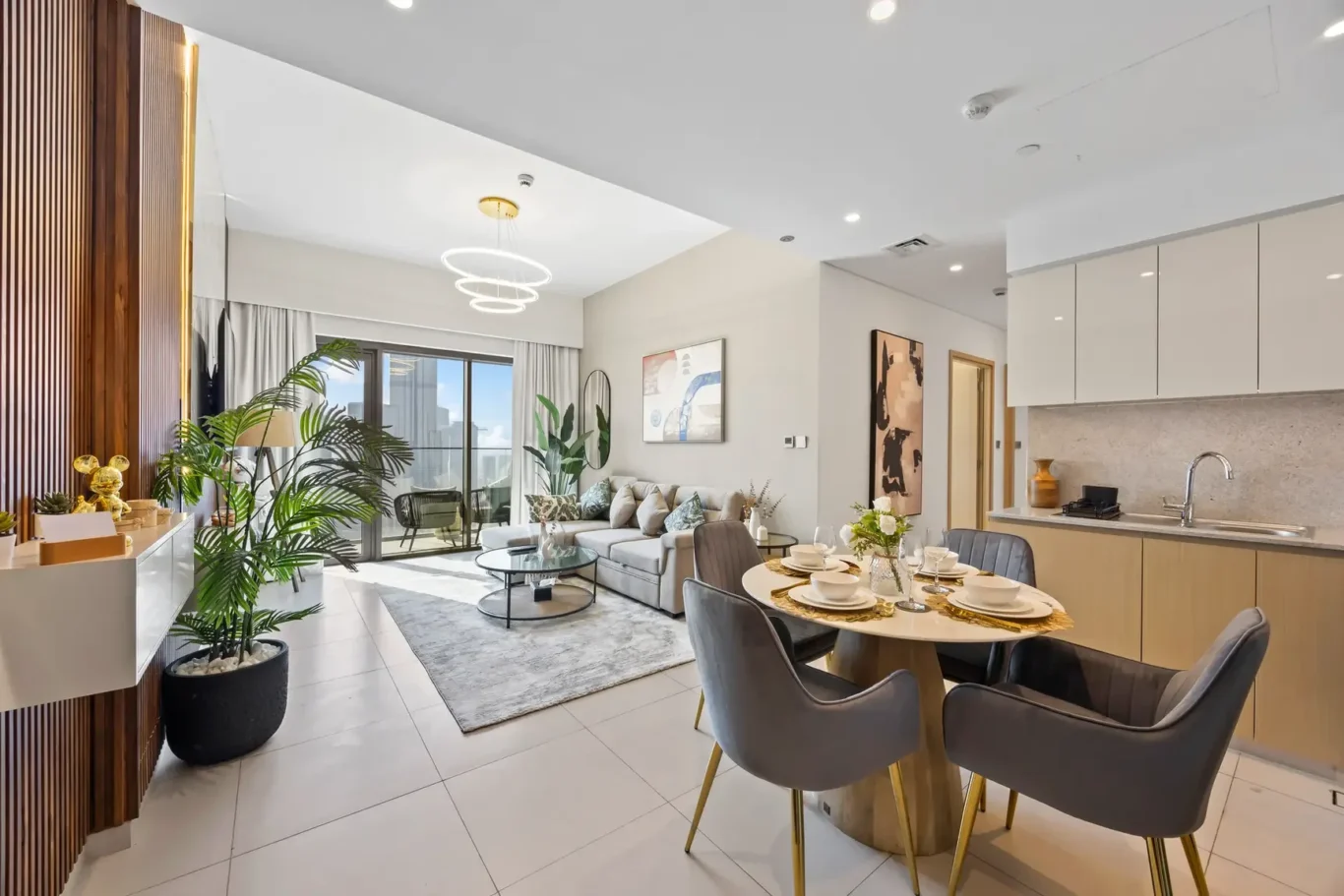 Open-concept living area with a gray sofa, glass coffee table, and large windows; dining table set for four near a modern kitchen.