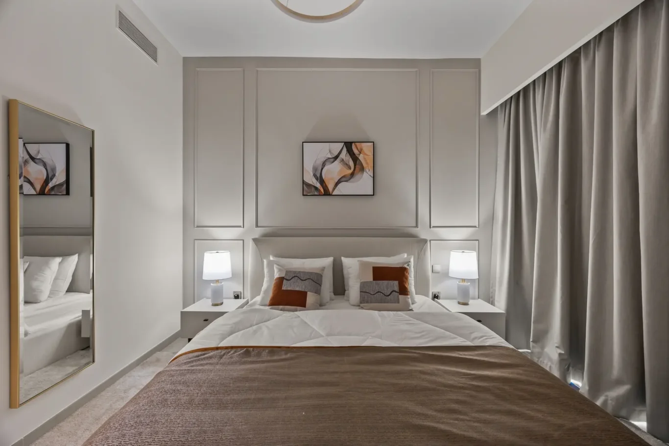 Modern hotel bedroom with a padded headboard, two white lamps on nightstands, gray paneled walls, and an abstract framed artwork above the bed is centered between pillows. The room also features heavy gray curtains.