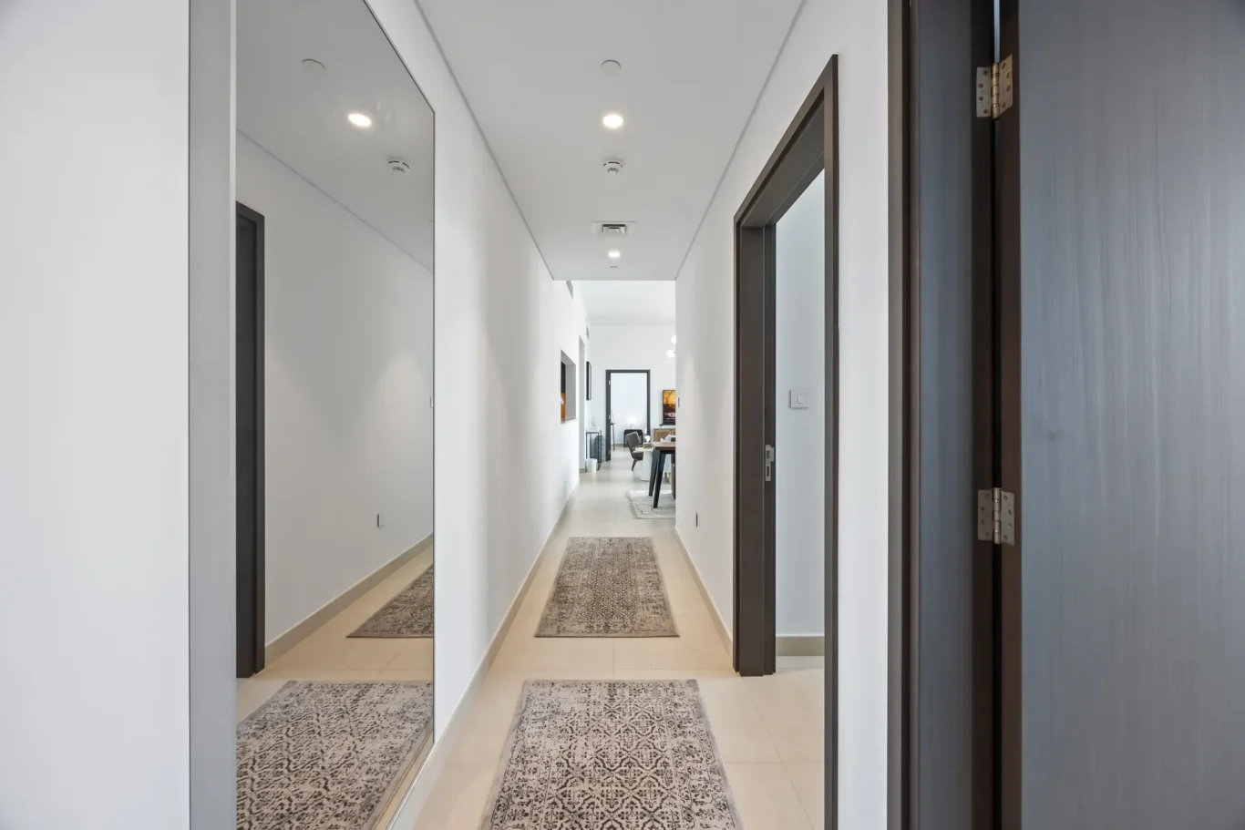 Narrow modern hallway with white walls, mirrored left wall, dark-framed doors, and a living area visible at the end.