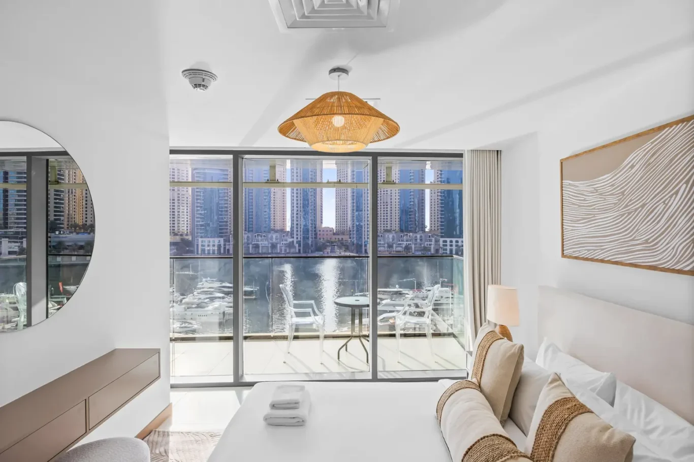 Bright hotel room with a large glass door balcony overlooking a marina and city skyline, with a bed in the foreground and neutral decor.