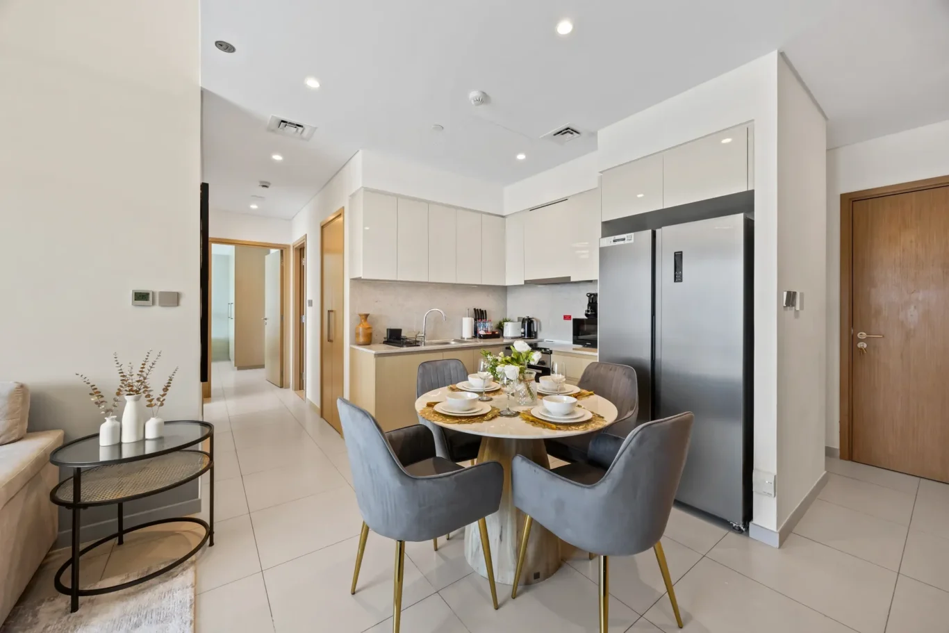 Bright open-plan kitchen with white cabinets, stainless fridge, and a round dining table set for six in the foreground.