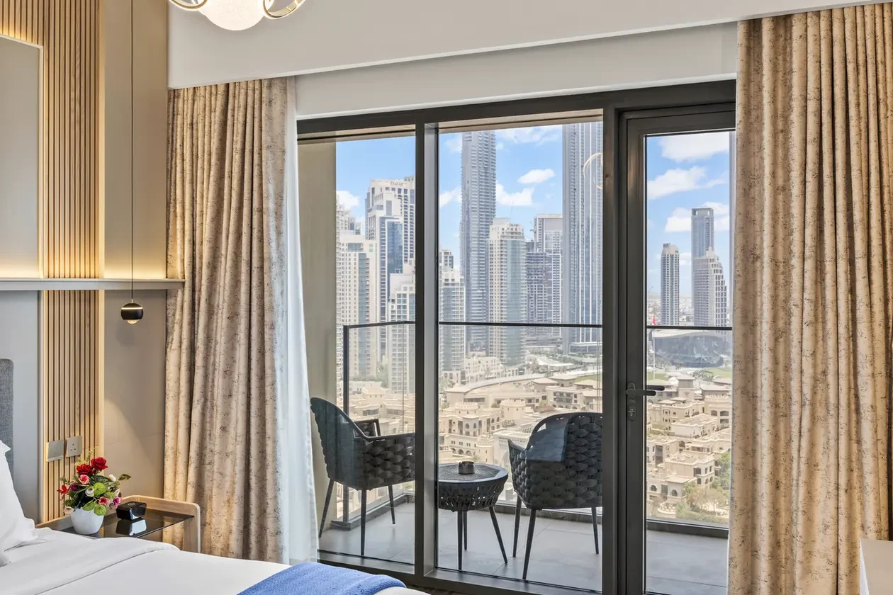 Hotel bedroom with glass doors opening to a balcony; city skyline with tall buildings outside, two chairs and a small table on the balcony.