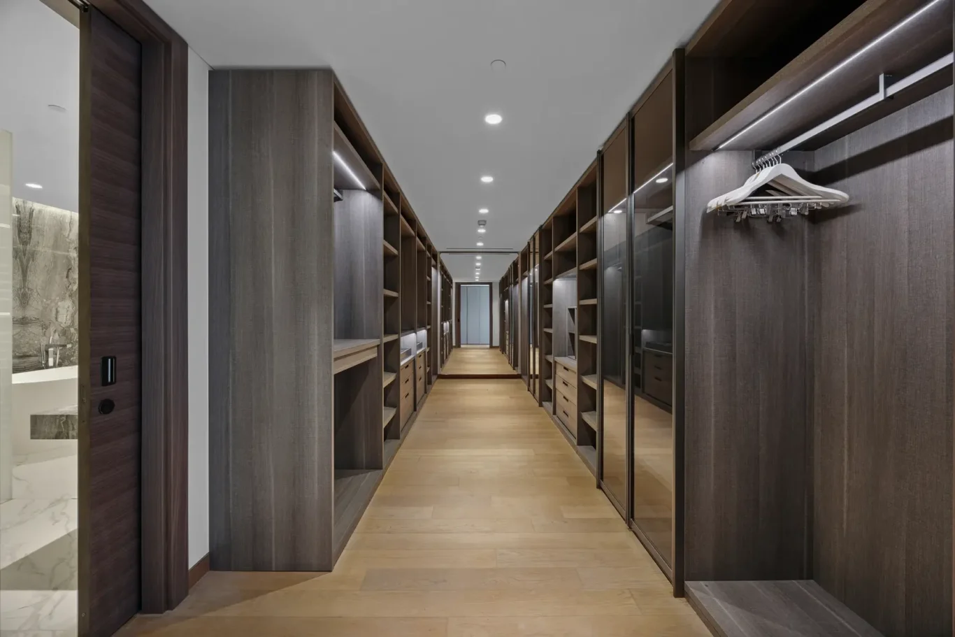 Wide walk-in closet with dark wood cabinetry on both sides, open shelves, hanging rods, and built-in drawers, lit by recessed ceiling lights.
