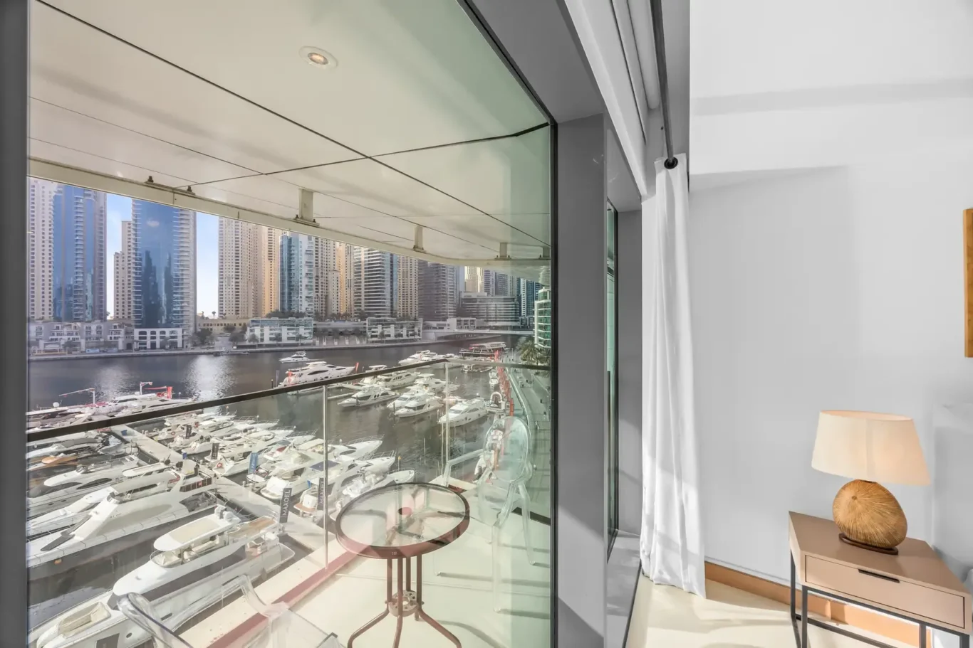 Modern living area with a glass wall overlooking a marina filled with yachts and a city skyline; white curtains, small round table and lamp nearby.