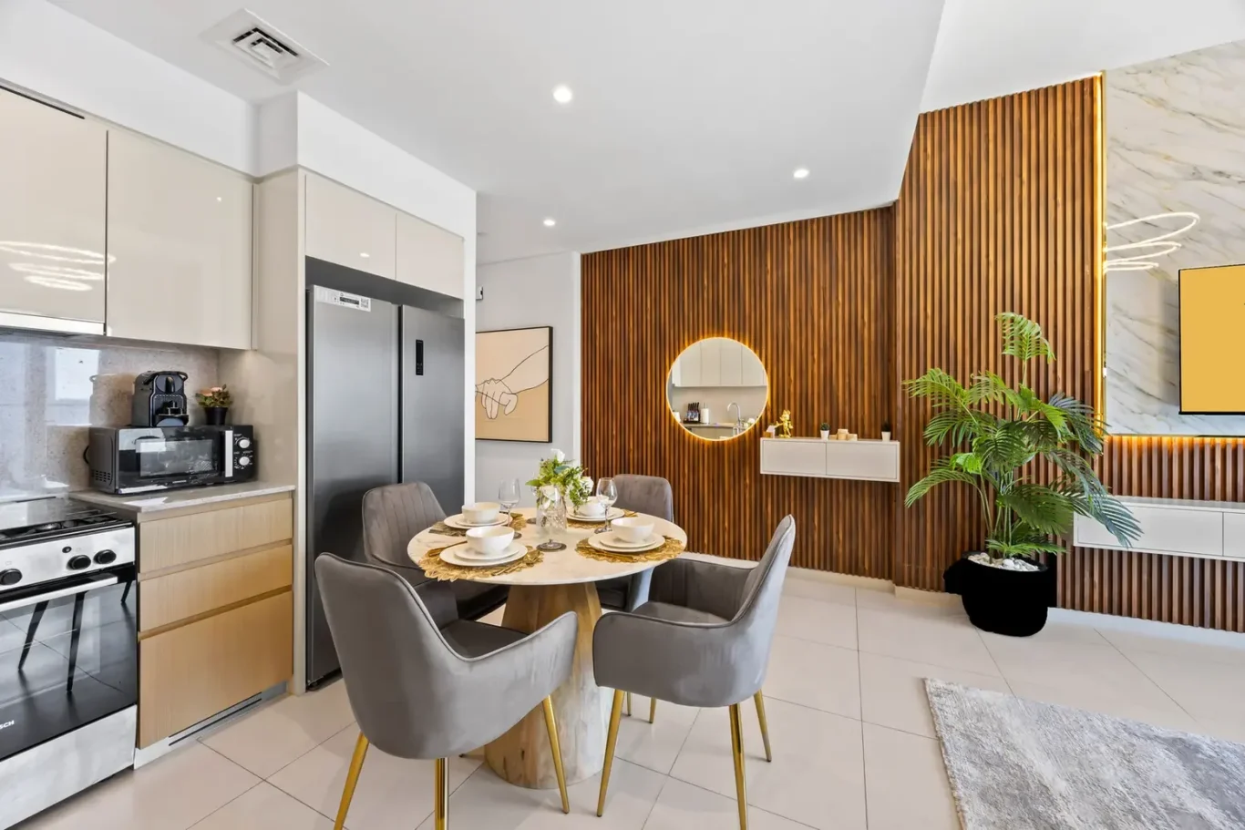 Open-plan kitchen and dining area featuring a round marble table set for four, gray upholstered chairs, and a vertical wood slat wall.
