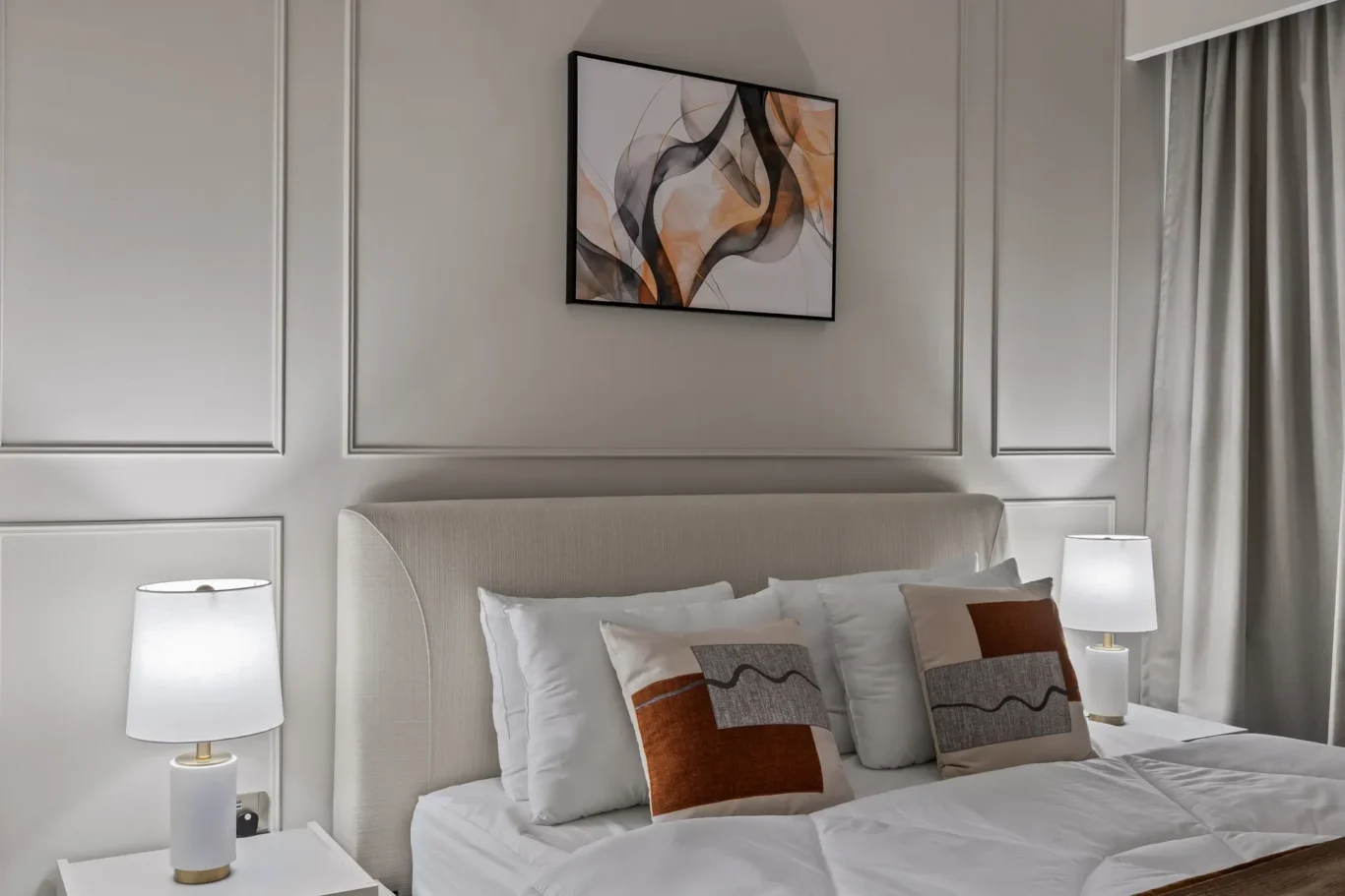 Hotel-style bedroom with a tufted beige headboard, framed abstract art, and two bedside lamps