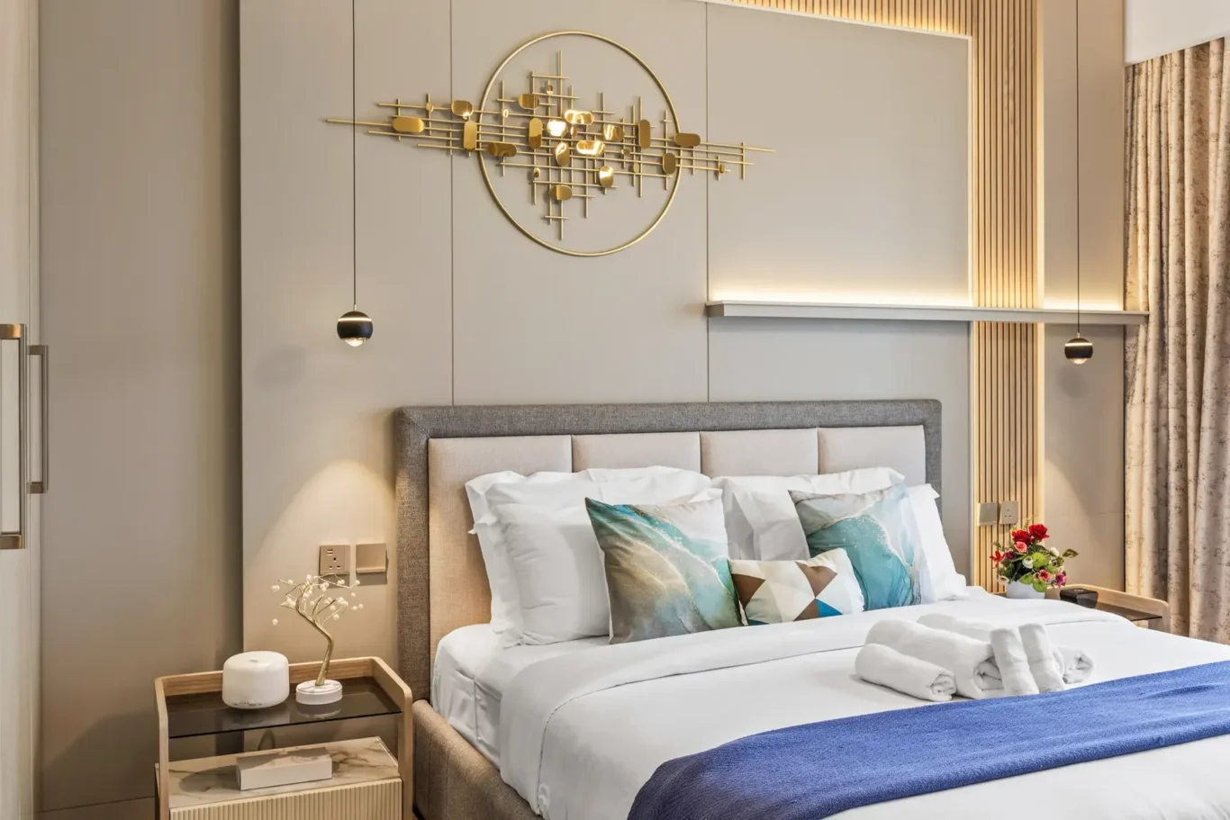 Hotel bedroom with an upholstered gray headboard, white bedding, and a blue throw at the foot of the bed; gold geometric wall art above.