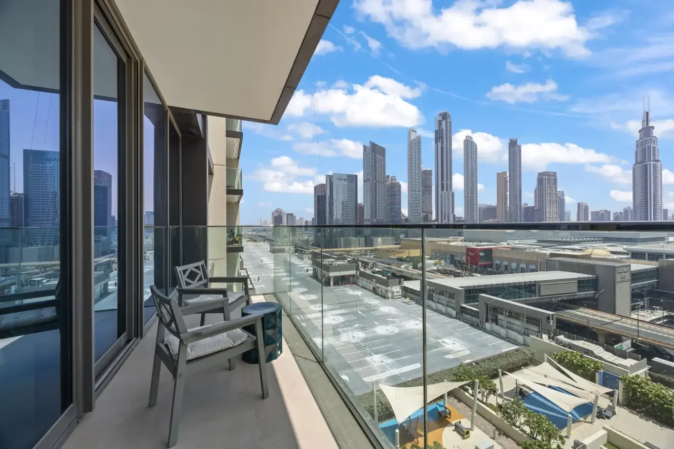 Balcony with two chairs and a glass railing overlooking a city skyline of tall skyscrapers under a blue, partly cloudy sky.