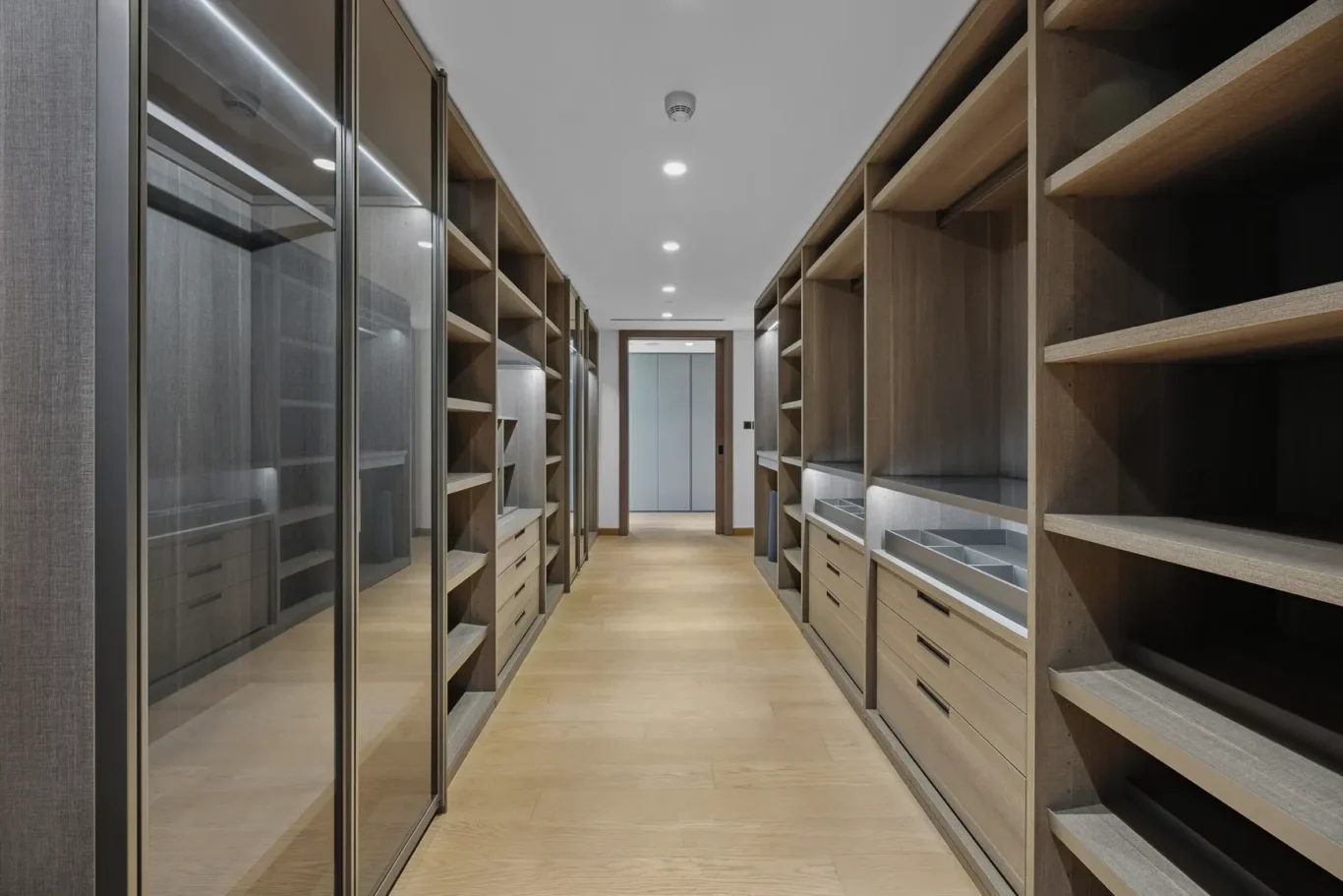 Modern walk-in closet with glass-front cabinets on the left and open wooden shelves on the right, illuminated by recessed ceiling lights.