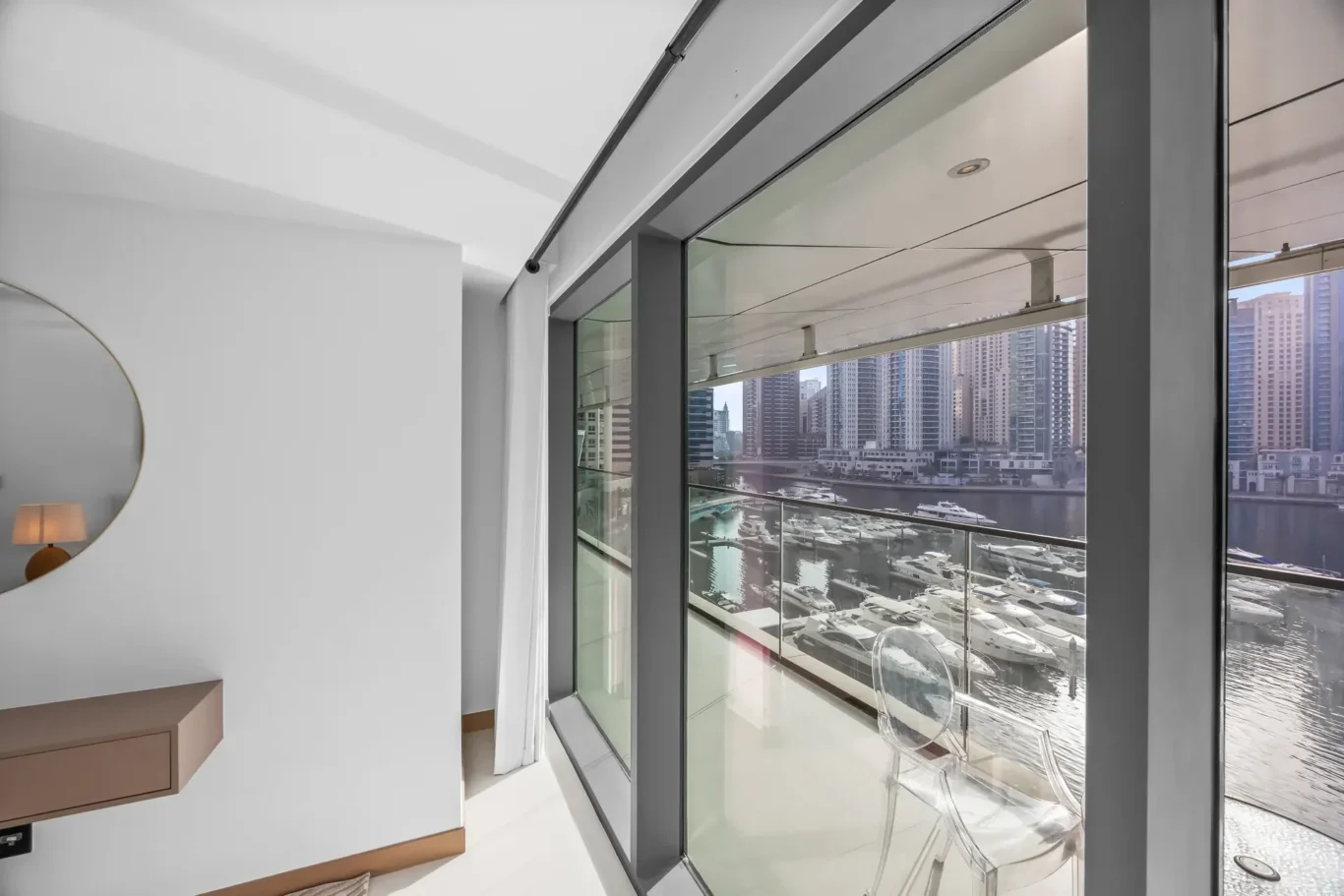 Bright modern room with floor-to-ceiling glass doors showing a marina and city skyline; round mirror and floating shelf visible on the white wall.