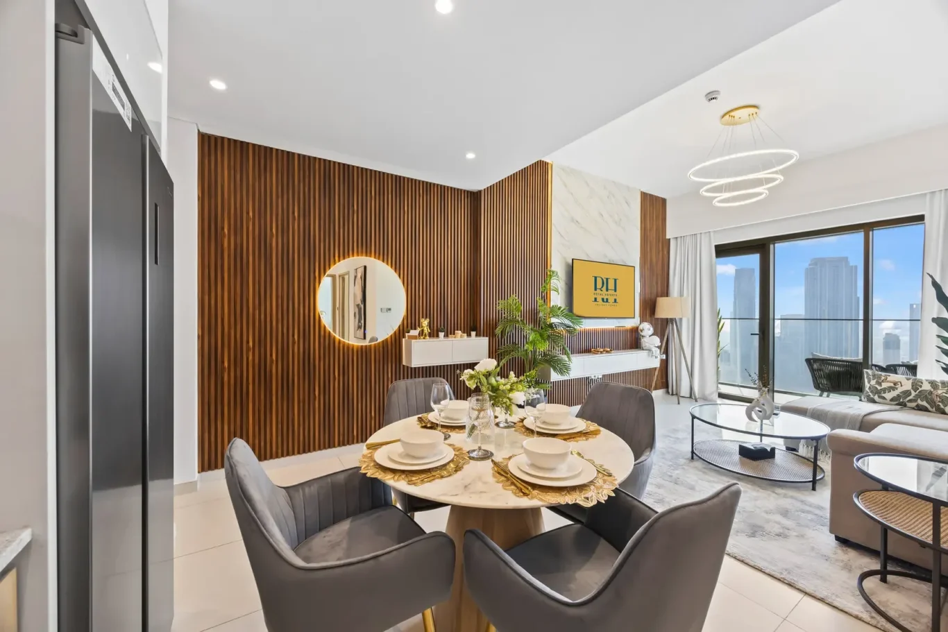 Open-plan dining area with a round marble table set for six, gray upholstered chairs, and a wood slat accent wall with a round mirror.