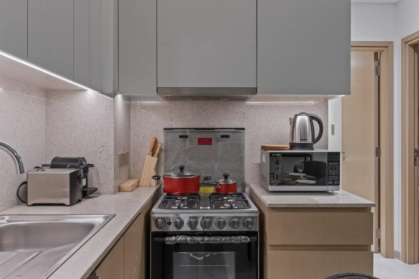 Modern compact kitchen with gray upper cabinets, beige lower cabinets, and a stainless steel gas range at center with red pots on the stove.