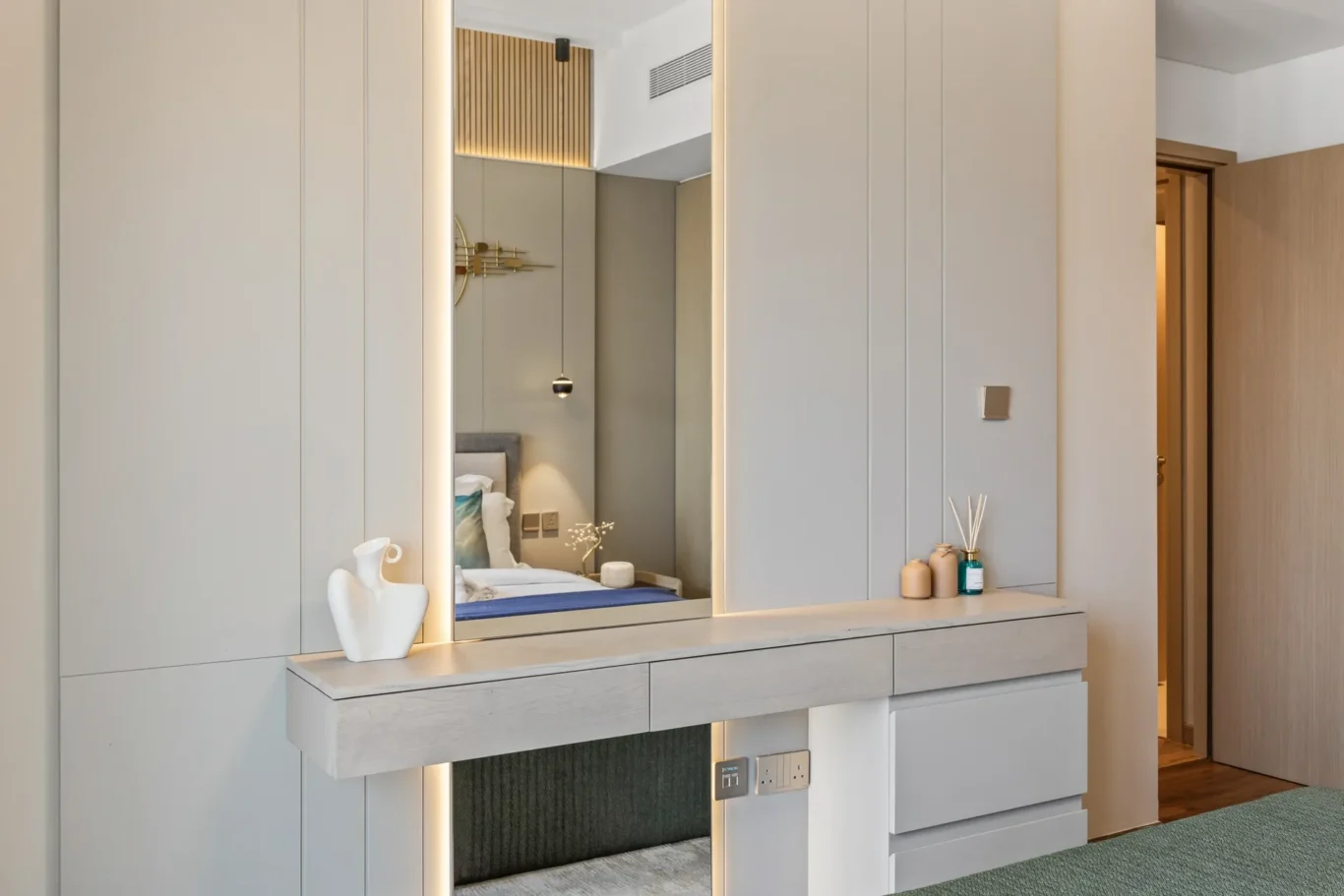 Modern bedroom vanity with a backlit framed mirror, beige cabinetry, and a floating drawer unit; decorative vases sit on the surface while the bed is visible in the mirror.