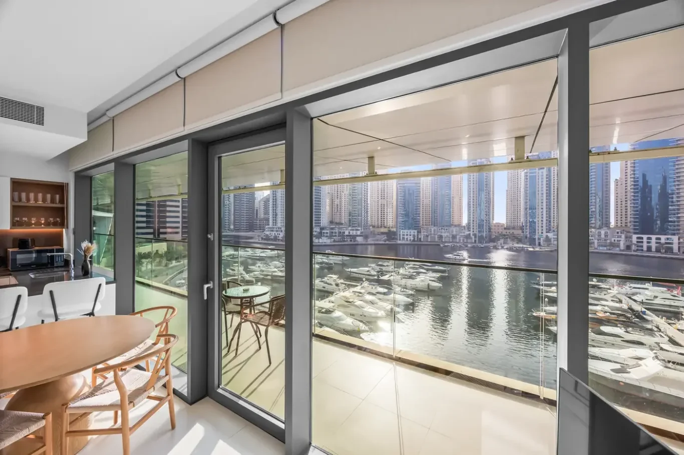 Modern dining area with a round wooden table and chairs, open to a glass balcony overlooking a marina with yachts and city high-rises in the distance.