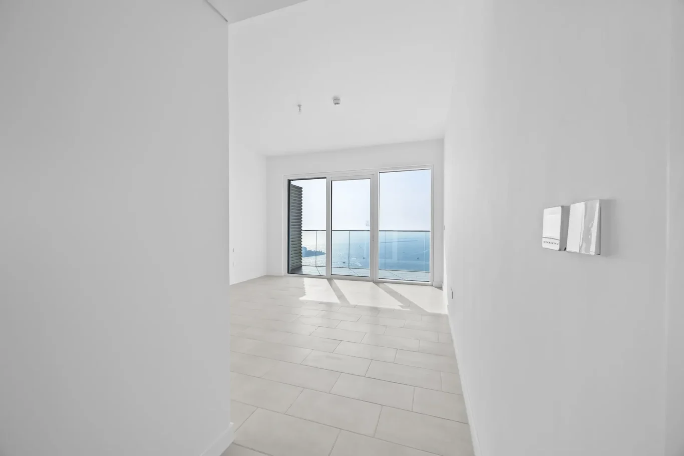 Empty white room with large sliding glass doors opening to a balcony overlooking the sea.