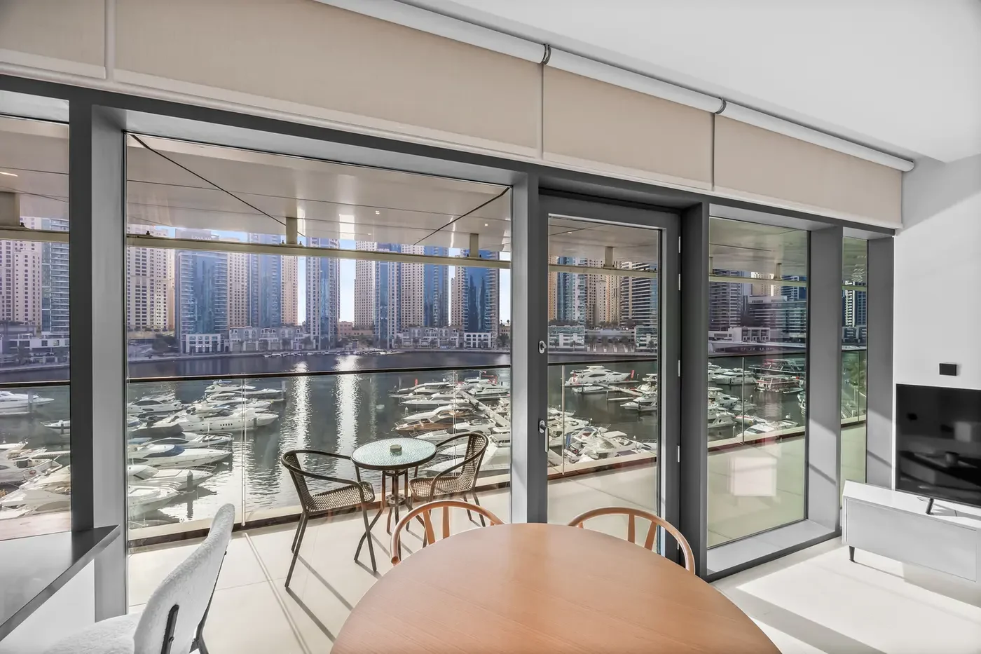 Modern dining area with a round wooden table overlooking a marina filled with yachts through glass doors.",