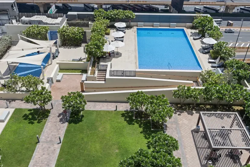 A rooftop pool area with a rectangular blue pool, white umbrellas, lounge chairs, and surrounding trees on a paved terrace.