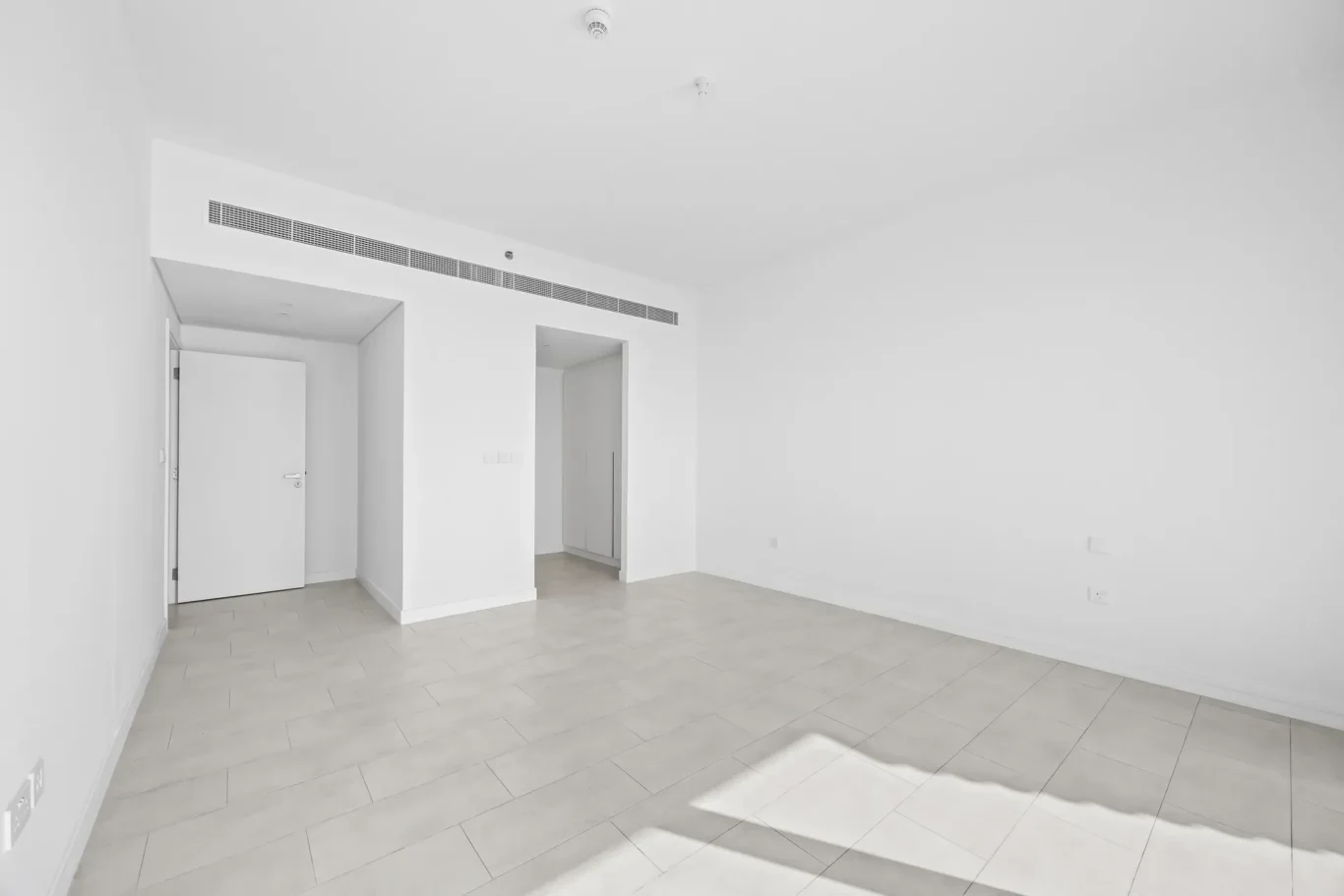 Bright empty white room with pale tiled floor, a door on the left, and an open passage leading to a back hallway or storage area.