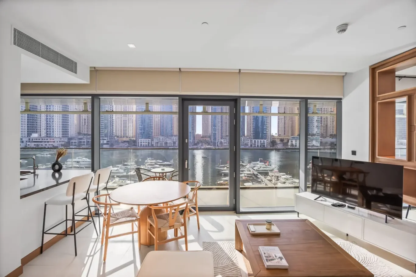 Bright living room with a round dining table, white chairs, and large windows showing a marina and city skyline.
