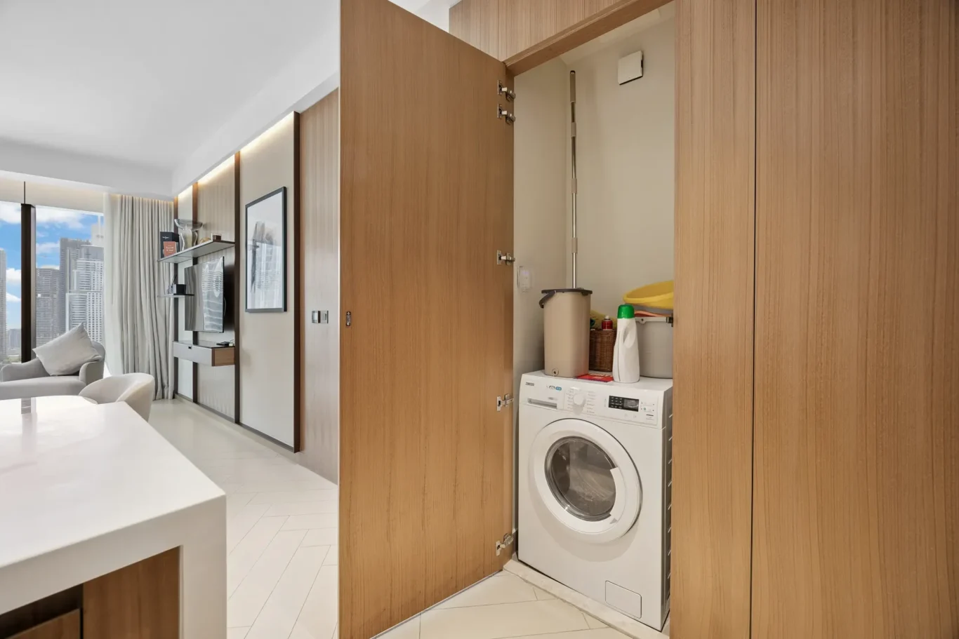 Wood-paneled laundry cabinet with doors open, showing a front-loading washing machine and detergent items inside. Adjoins a bright living area with large windows.