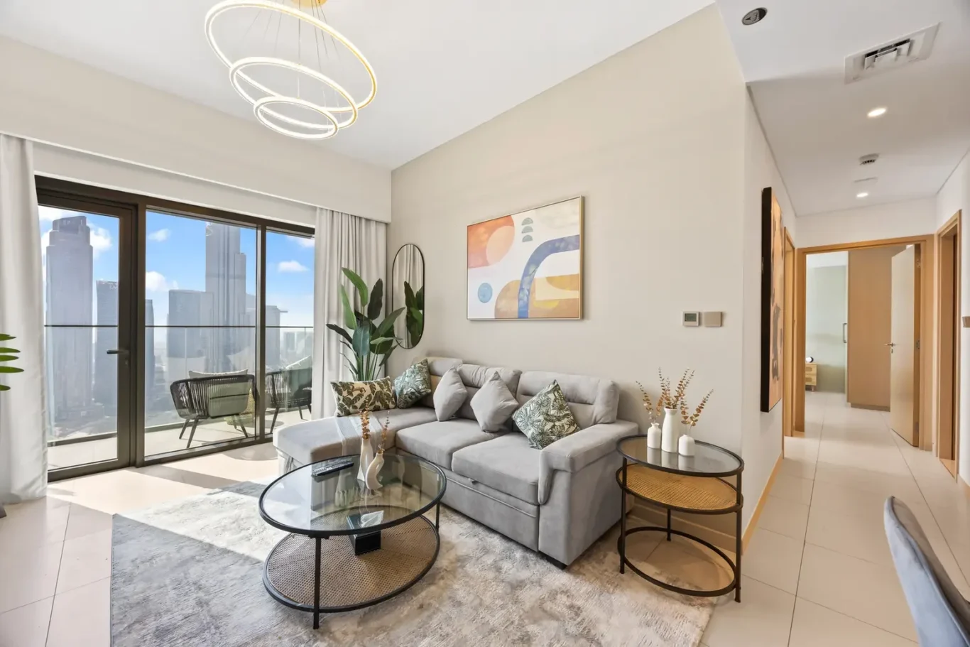 Bright modern living room with a light gray sectional, abstract art, and two glass coffee tables overlooking a city skyline through large sliding glass doors.