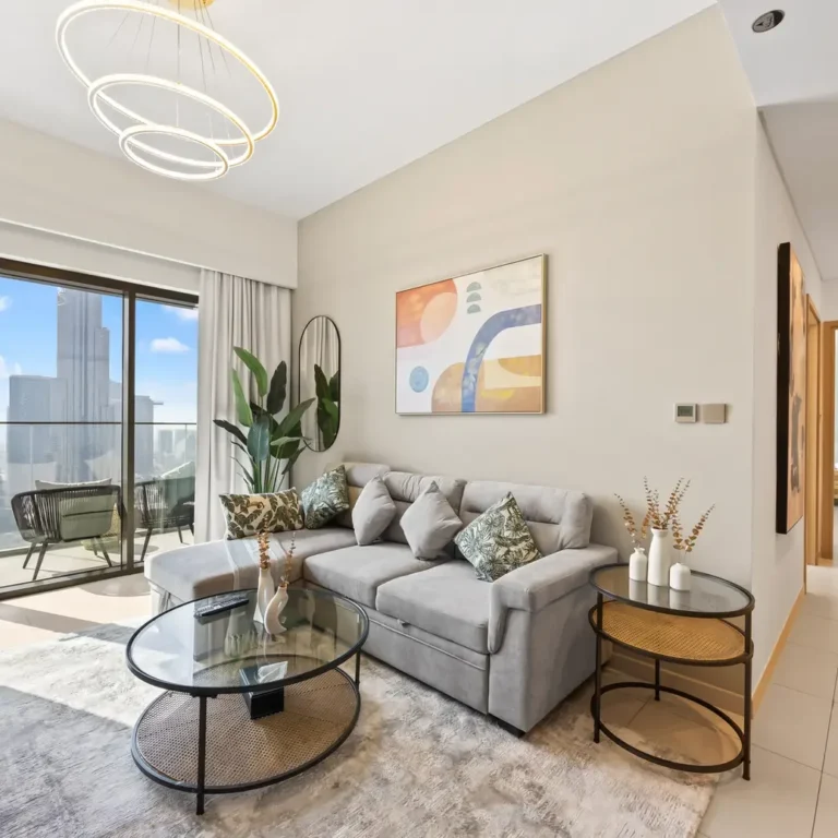 Bright modern living room with a light gray sectional, abstract art, and two glass coffee tables overlooking a city skyline through large sliding glass doors.