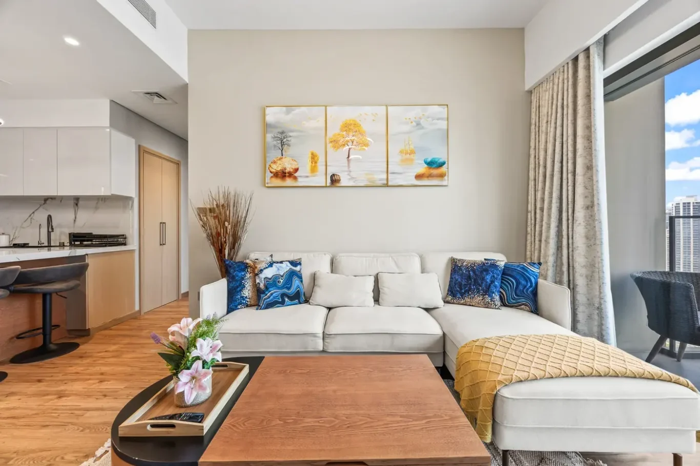 Bright open-plan living room with white sectional sofa, blue patterned cushions, and a mustard throw. A triptych painting hangs on a beige wall above the sofa. A wooden coffee table with a vase of pink flowers sits in front; balcony door to the right shows city skyline.