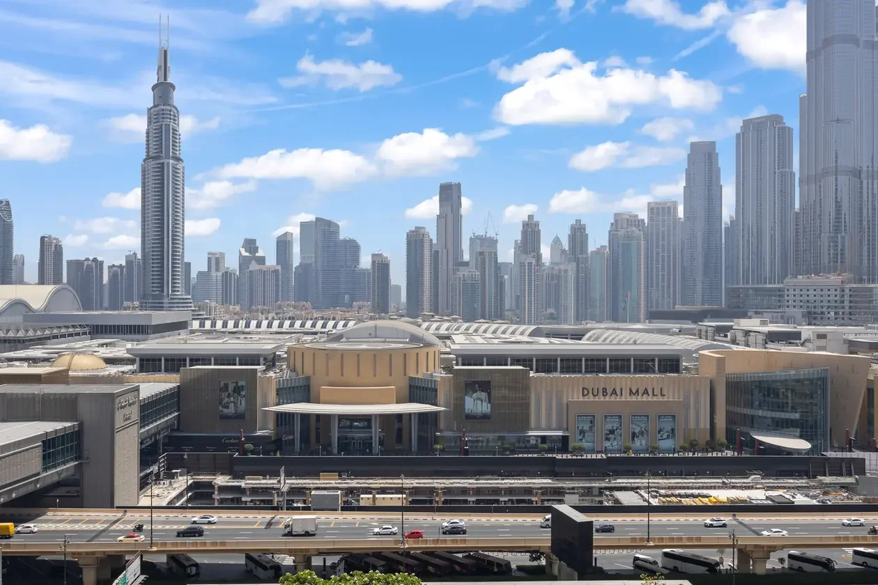 Dubai Mall entrance in the foreground with Burj Khalifa and a cluster of tall Dubai skyscrapers in the background under a blue sky.
