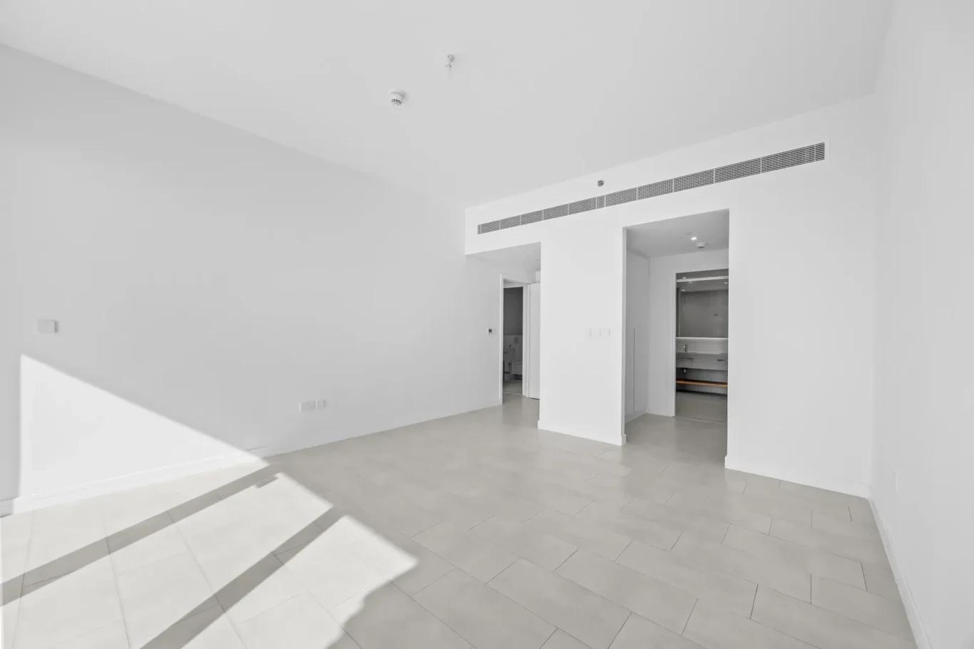 Bright, empty white living space with tiled floor; sun casts long shadows across the floor, and an open doorway to a kitchen area.