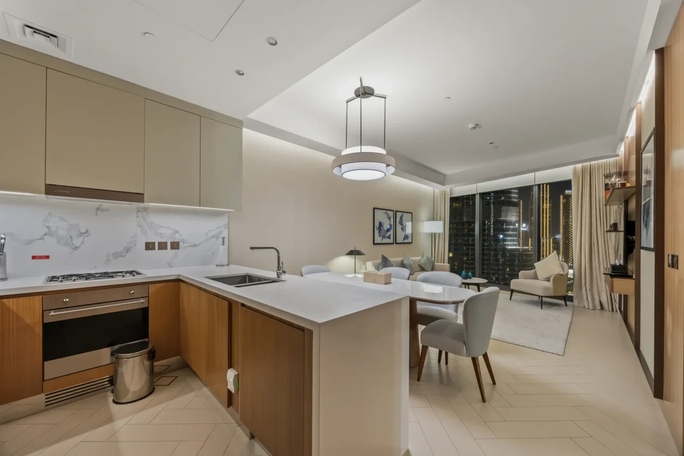 Open-concept kitchen with a white island, beige cabinets, marble backsplash, and a living area with city lights outside the windows at night.