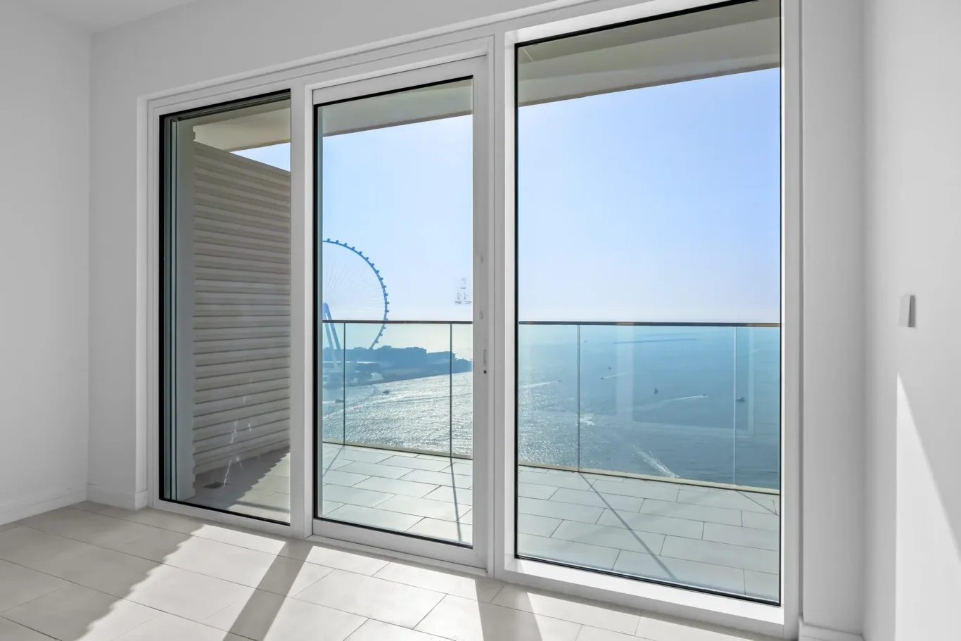 Interior space with large glass sliding doors opening to a balcony, offering a bright ocean view and a distant Ferris wheel outside.
