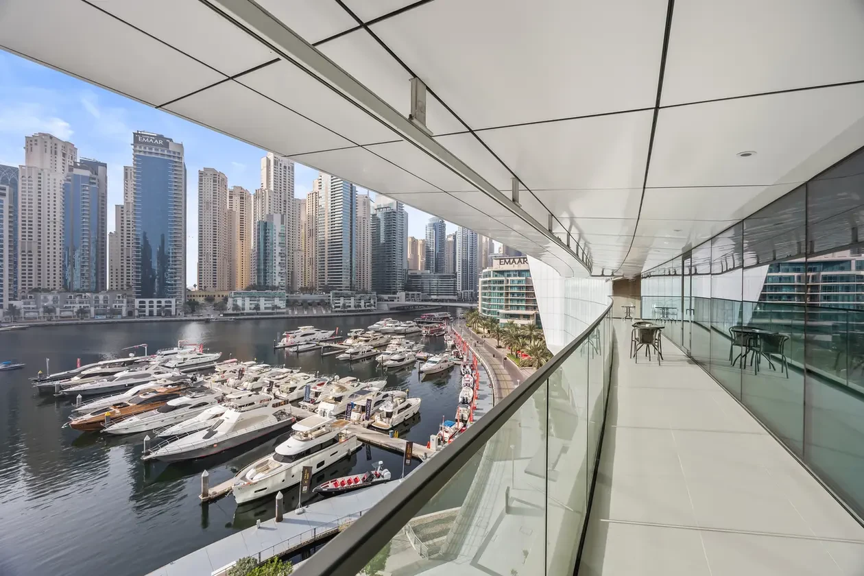 Marina filled with luxury yachts docked along the water, with a curved glass-walled promenade and a skyline of tall modern towers in the background.
