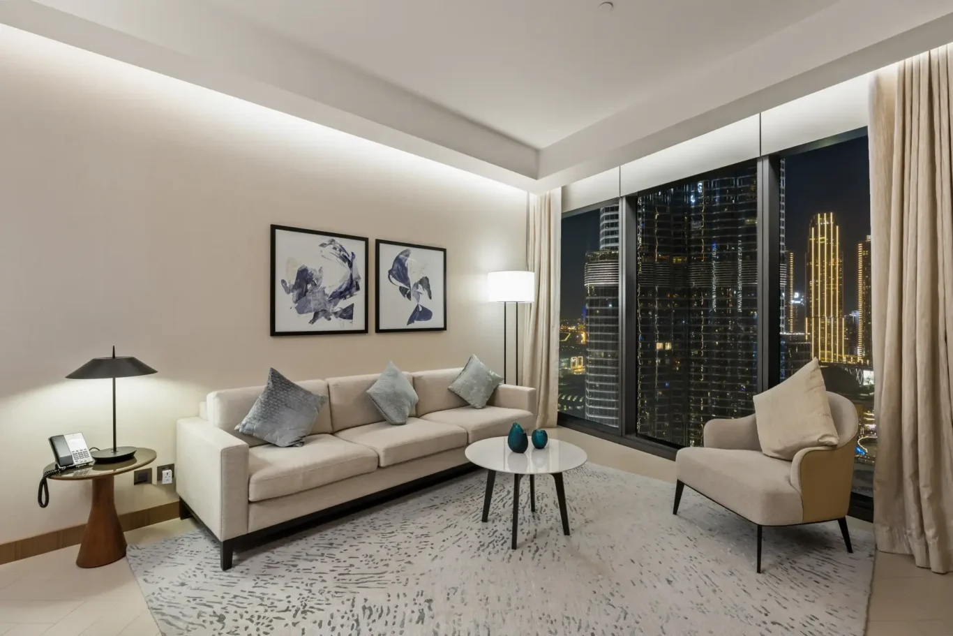 Hotel living room at night featuring a beige sofa with throw pillows, round coffee table, floor lamp, abstract art, and floor-to-ceiling windows overlooking a lit city skyline.