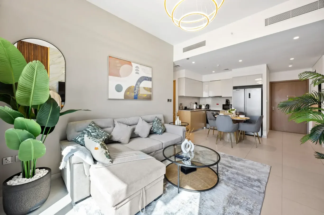 Open-plan living area with a grey sectional sofa, abstract art, large green plants, and a glass coffee table, connected to a modern kitchen and dining space.