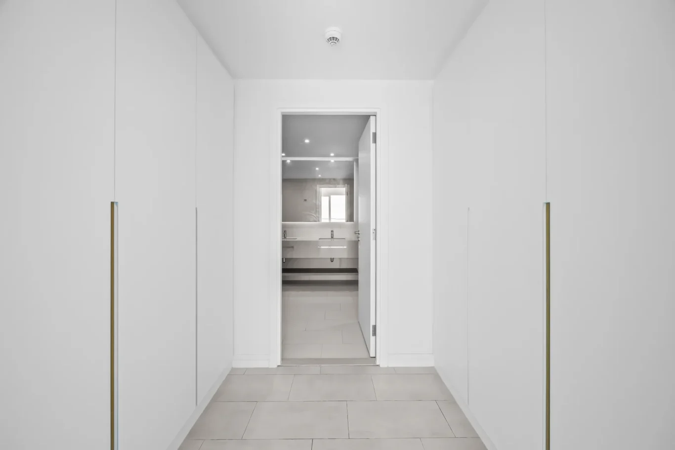 White hallway lined with tall white built-in cabinets with gold handles, opening to a bright bathroom at the end.