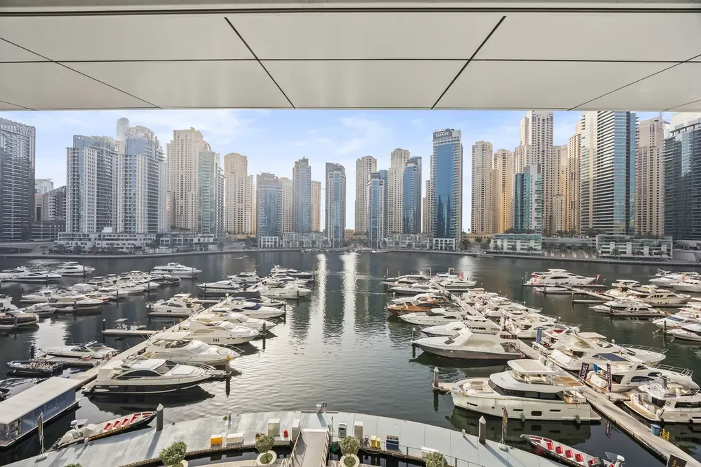 Marina filled with luxury yachts docked, with a dense row of glass skyscrapers in the background under a blue sky.