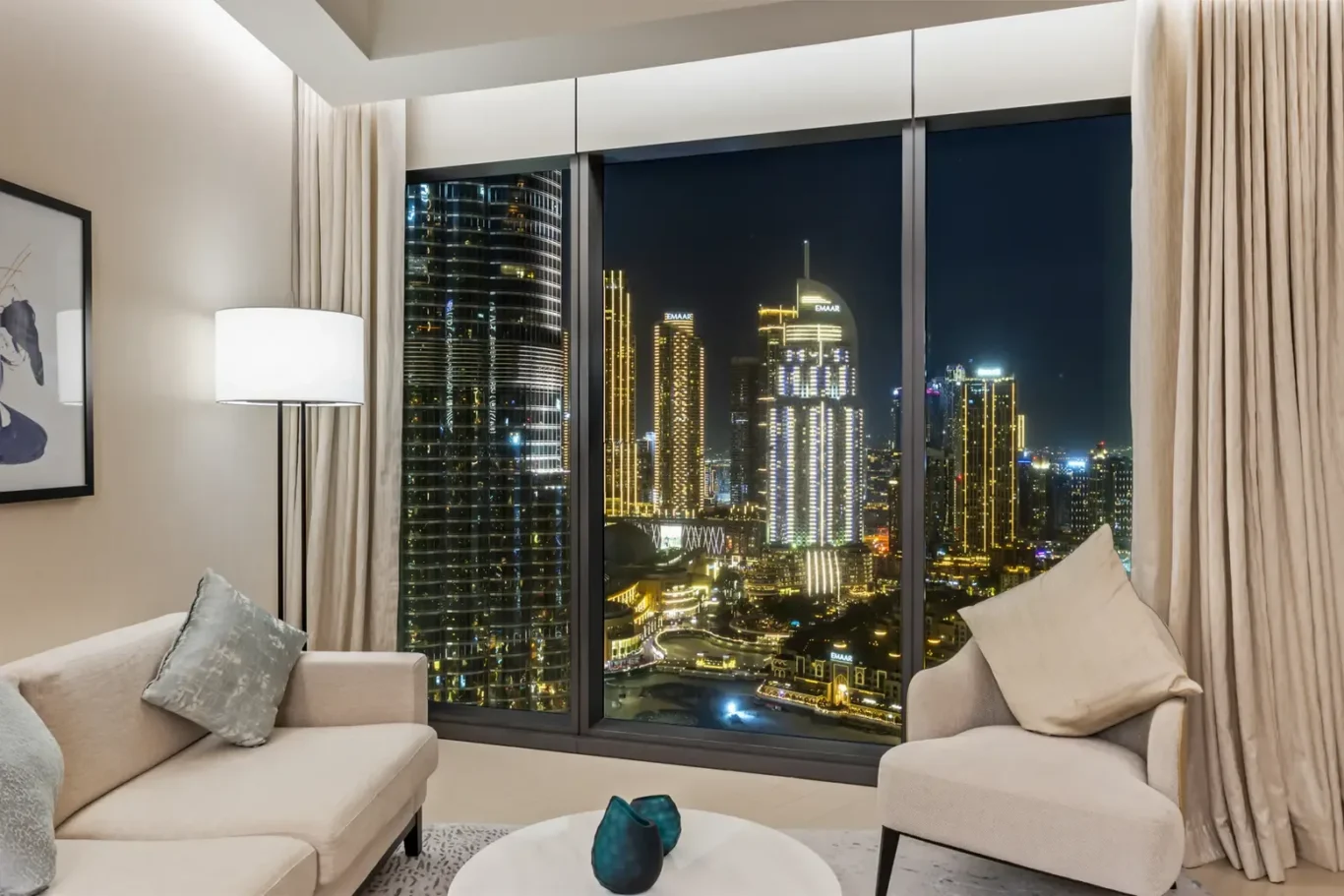 Living room at night with a city skyline visible through large floor-to-ceiling windows, beige sofa and armchair.