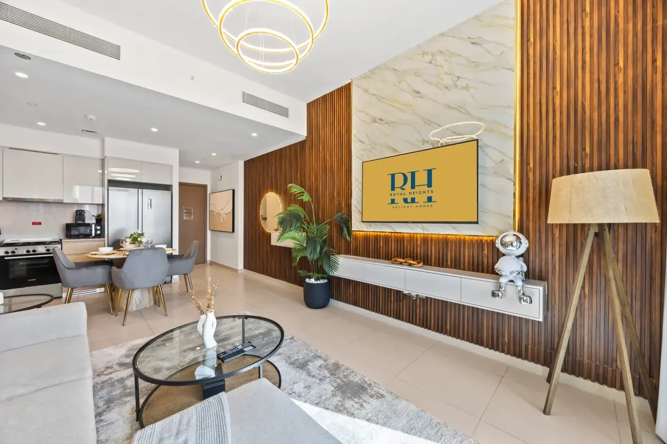 Open-concept living area with a grey sofa, glass coffee table, and a dining table in a bright kitchen, featuring a wood slat wall and modern circular pendant lights.