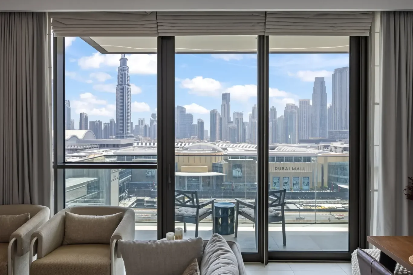Living room with a balcony view of Dubai's skyline through large glass doors and a mall below.