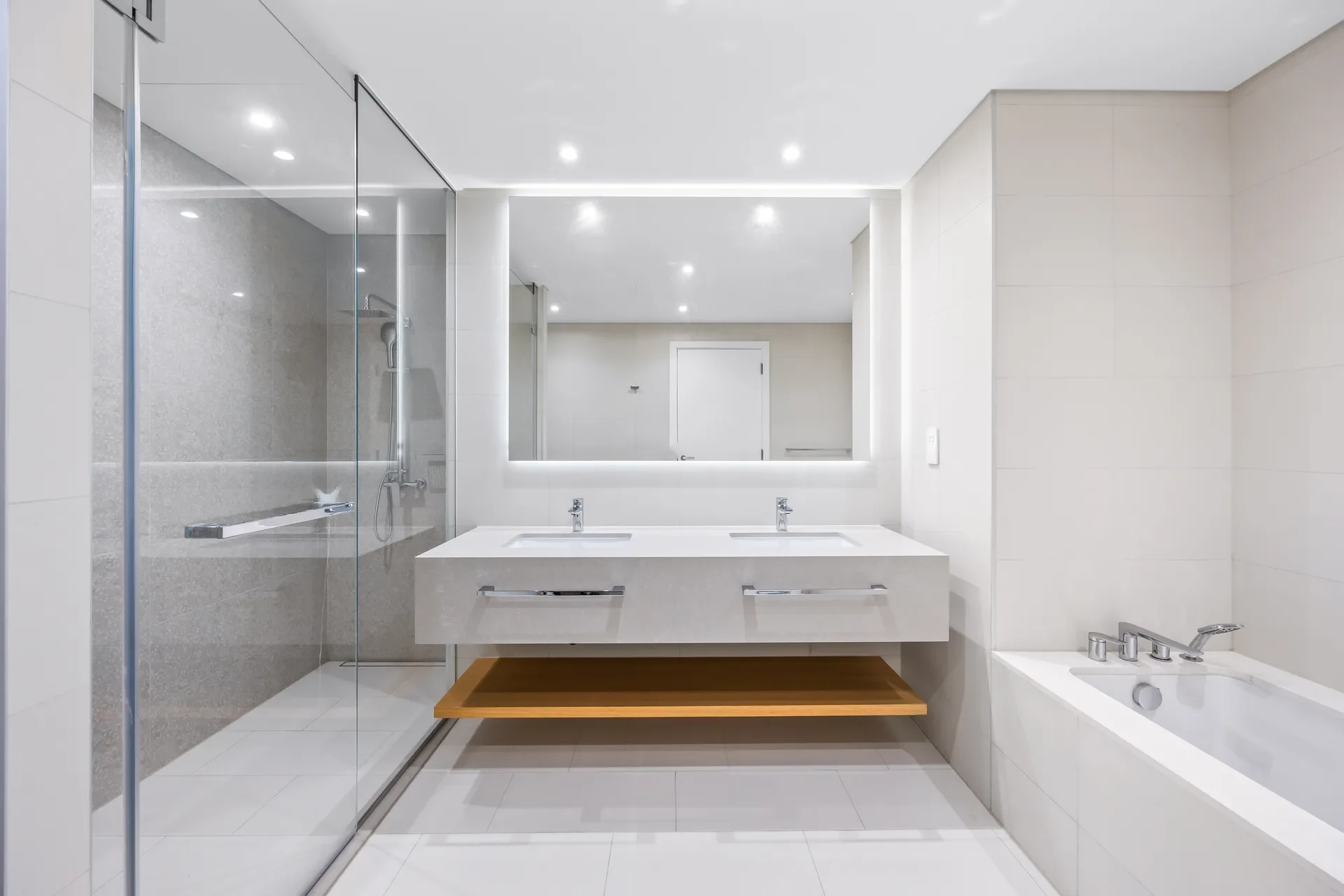 Modern bathroom with double sinks and backlit rectangular mirror, glass-enclosed shower on the left and a bathtub on the right.