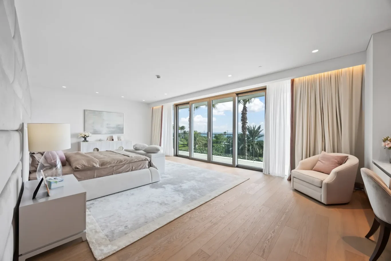 Bright hotel bedroom with a large glass wall opening to a balcony, overlooking palm trees and the sea.