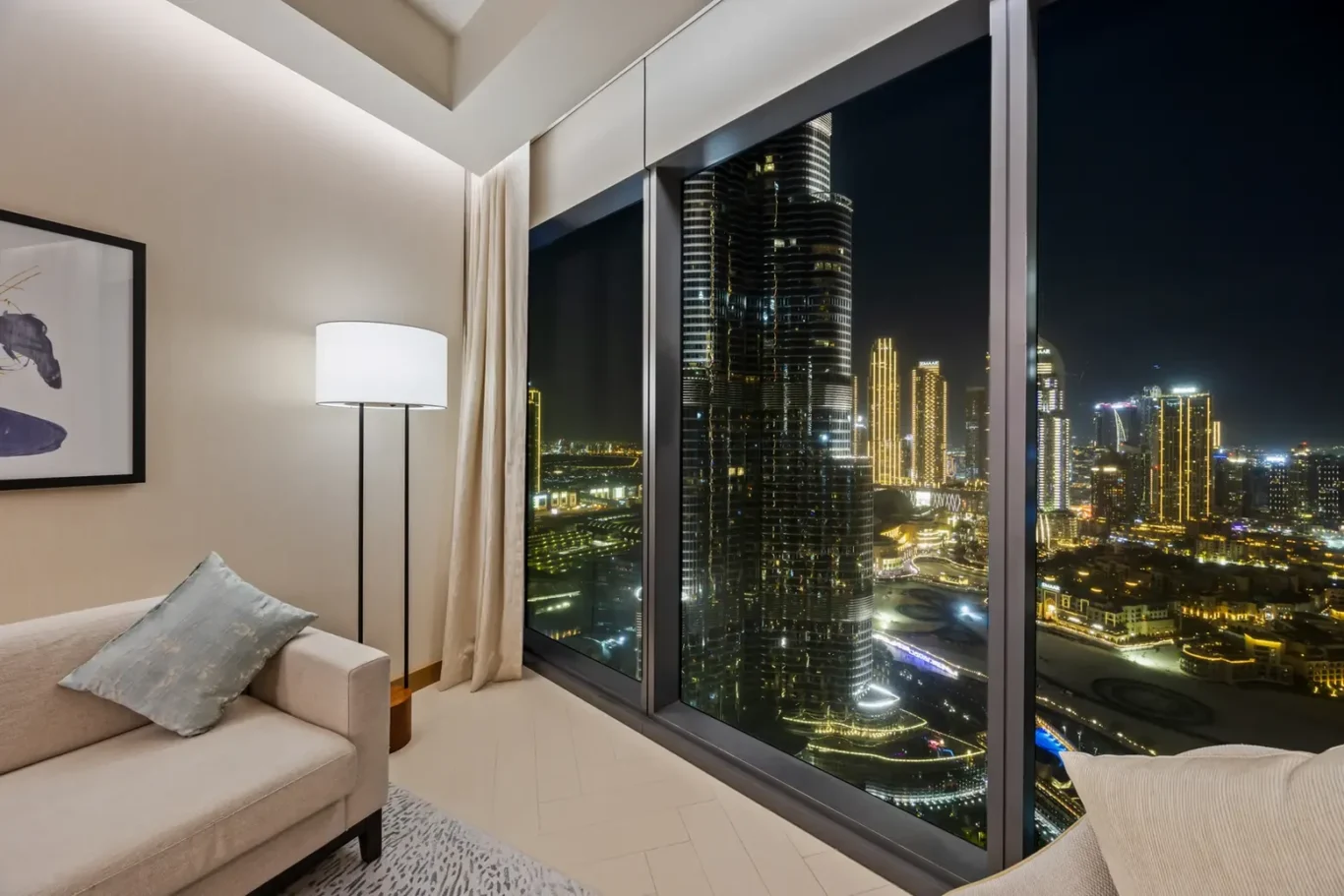 Night city skyline visible through floor-to-ceiling windows from a modern living room with illuminated skyscrapers outside.
