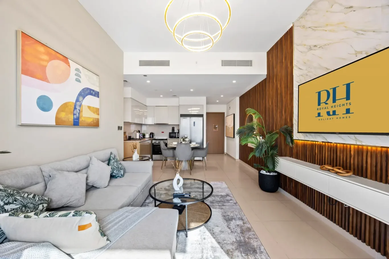Open-concept living room with gray sectional, round glass coffee table, and abstract wall art; kitchen in back and pendant rings overhead.