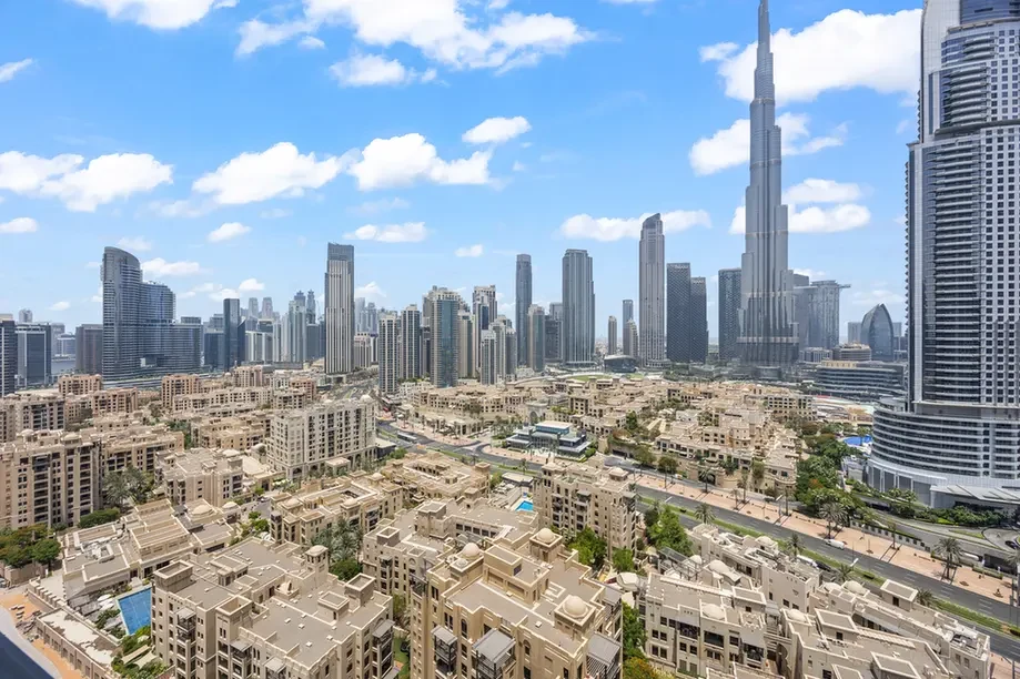 Wide cityscape with a dense cluster of beige low-rise homes in the foreground and towering glass skyscrapers under a blue sky with scattered clouds, likely a modern urban center.